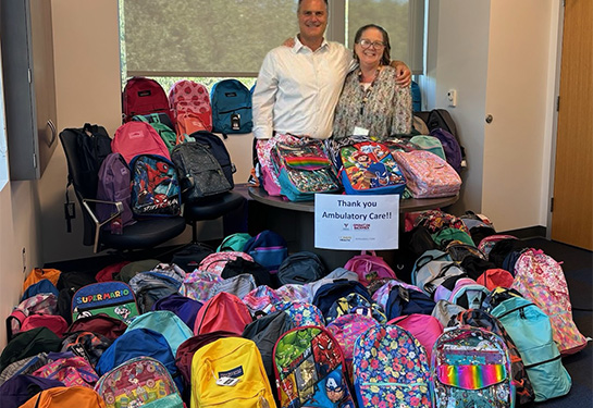 Two adults stand amid a pile of colorful backpacks