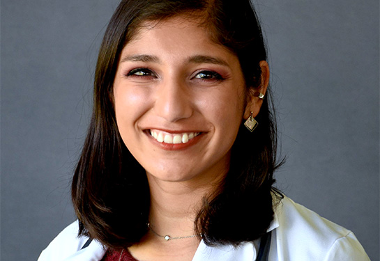 A young woman with dark hair past her shoulders, red wine-colored top and white lab coat, and stethoscope around her neck