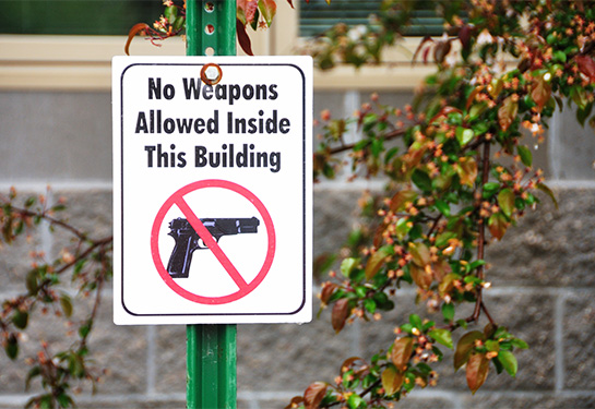 A sign that reads &#x201c;no weapons allowed&#x201d; with a gun with a red line through it is nailed to a post near a tree. 