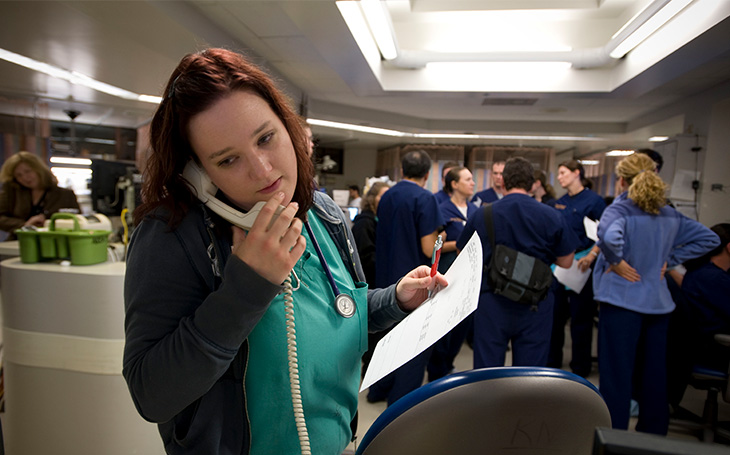 A nurse wearing green scrubs and talking on the phone with a care team standing behind her in the Emergency Department