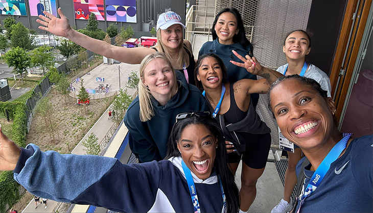 Marcia Faustin is taking a selfie with U.S. Gymnasts Simone Biles, Suni Lee, Jordan Chiles, Jade Carey and Hezly Rivera and coach Cecile Landi. They are standing on a balcony of their room in the Olympics Village in Paris.