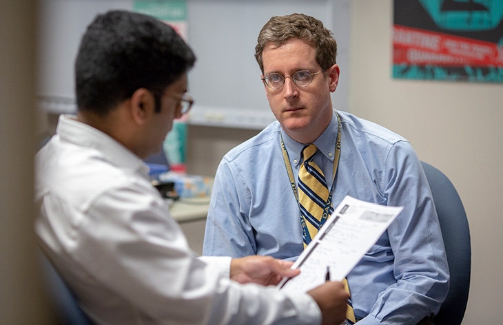 Dr. Stephen Henry wearing a blue long sleeve short and yellow necktie and looking at an individual wearing a white short and holding a paper and pen. Both individuals are wearing eyeglasses.