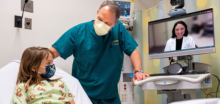 A health care provider talks to young patient at beside. A physician on telehealth interacts with them through a computer screen