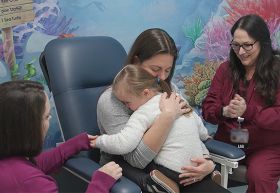 An adult sits in a medical chair with a child on their lap as a nurse begins a blood draw procedure and a second provider stands nearby.