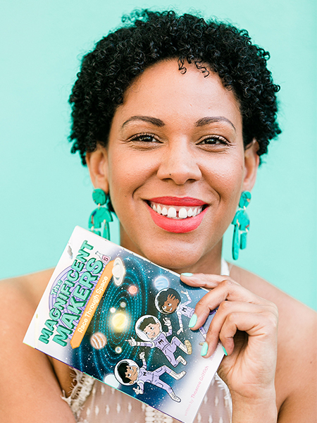 Woman with dark curly hair, green earrings and wearing a white dress holds a paperback book with the title, 