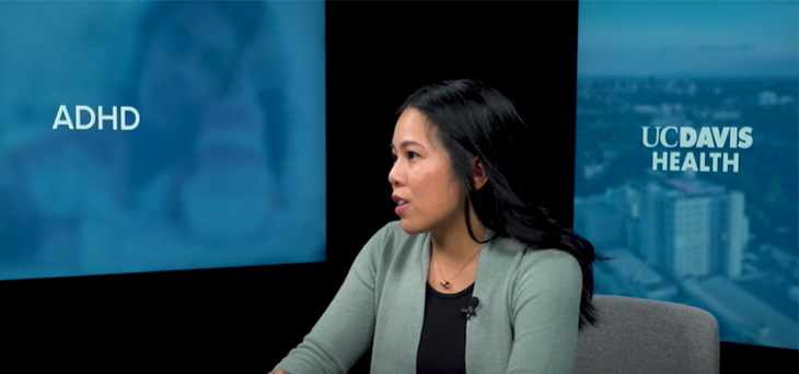 A woman with long dark hair sits at a table in front of two monitors with text 