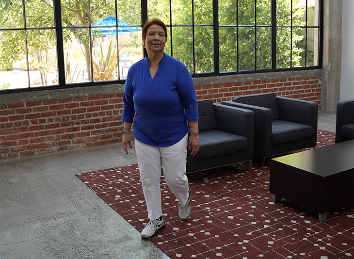 A patient in a bright blue shirt and white pants walks in a medical clinic with a concrete floor and maroon rug.