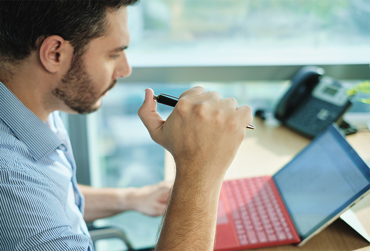 A man sits at a table, working on a laptop while holding a pen that he is clicking open in one hand.