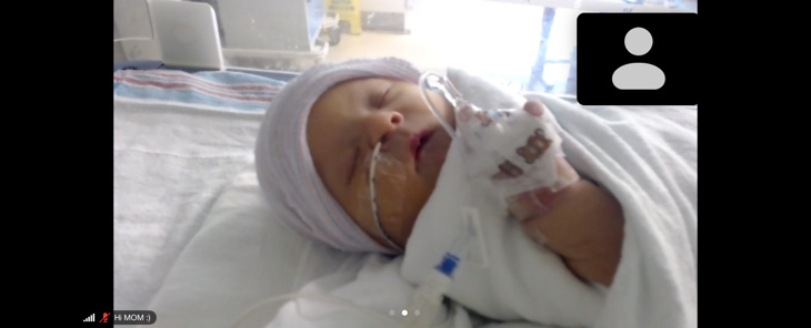 Baby with hat on and tubes in her nose wrapped in a blanket lying in a NICU bed.