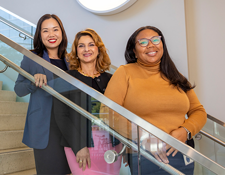 Three women stand on different steps of a stairwell in front of a glass railing