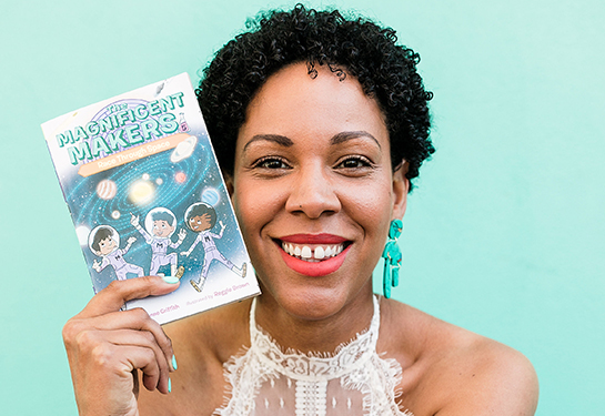 Woman with dark curly hair, green earrings and wearing a white dress holds a paperback book with the title, “Magnificent Makers.”