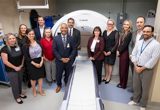 Group of people standing around a computed tomography scanner with a large donut shaped circle of the head of a bed.