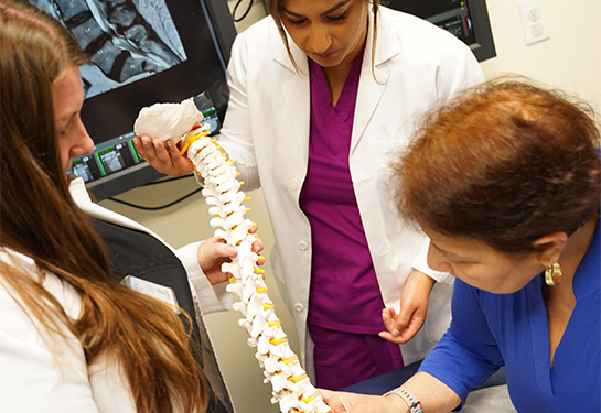 Two medical providers in white coats hold a model of a spine while meeting with a patient in a medical exam room. 