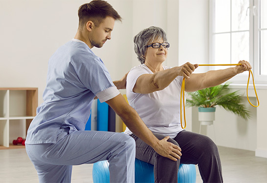 Male employee helps female senior patient with a physiotherapy exercise on fit ball. Woman on fit ball exercises with rubber band.