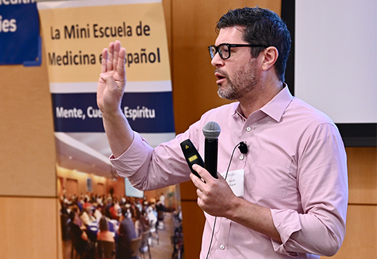 Un hombre de anteojos con camisa rosa sostiene el micrófono en su mano izquierda mientras habla frente a la audiencia