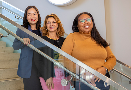 Three women stand on different steps of a stairwell in front of a glass railing