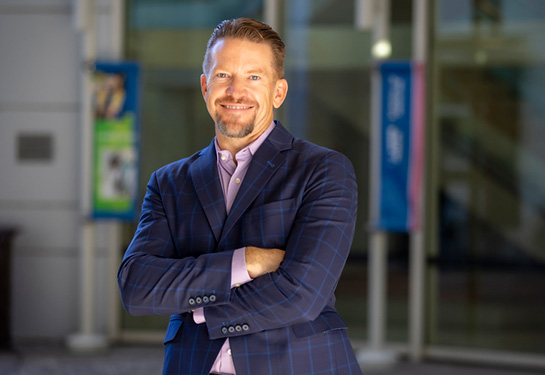 Dr. Nathaen Weitzel, wearing a blue suit with a lavender shirt, stands in front of glass doors on UC Davis Health&#x2019;s Sacramento campus.