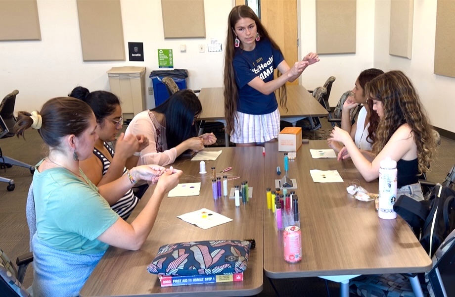 A young woman in a blue T-shirt that reads, “I am Tribal Health PRIME” holds a thread at a table where five students learn to bead