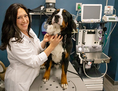 Woman in white coat smiling into camera with arm around large black/brown dog.