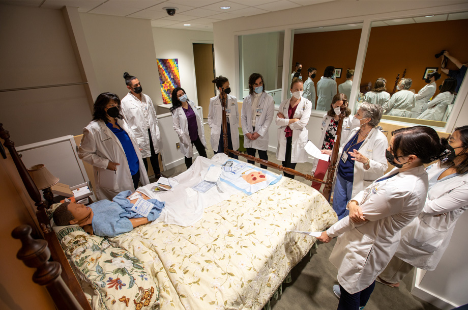 APP fellows wearing white coats and masks surround a bed with manikin laying in it and wound dressings on top of covers