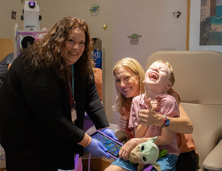 A mom sits with her young son in a medical chair while a child life specialist looks on nearby. All three are smiling widely. 