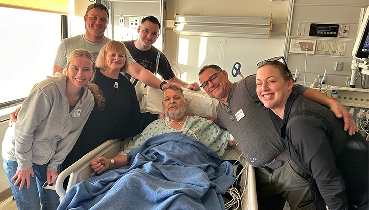 Six smiling family members stand around a hospital bed where an older male patient rests.