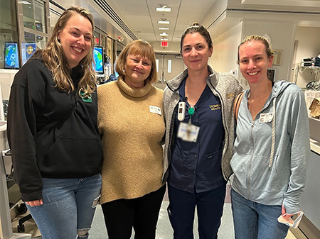 Four smiling women stand closely in a hospital ICU hallway.