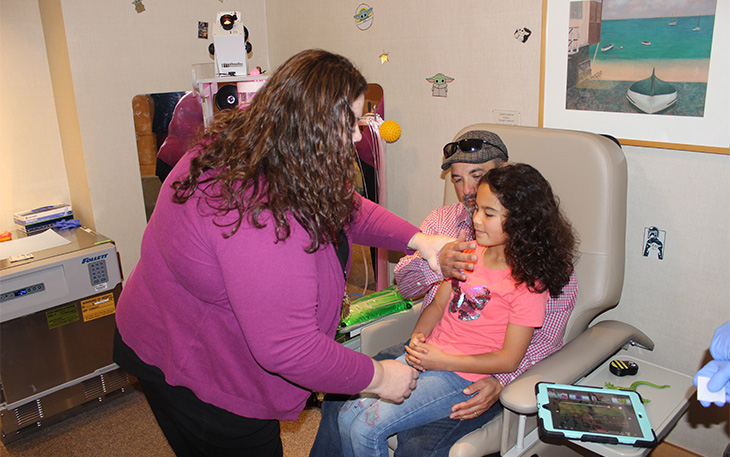 A child life specialist shows a father whose young daughter is sitting on his lap, how to hold her comfortably to receive a flu shot.