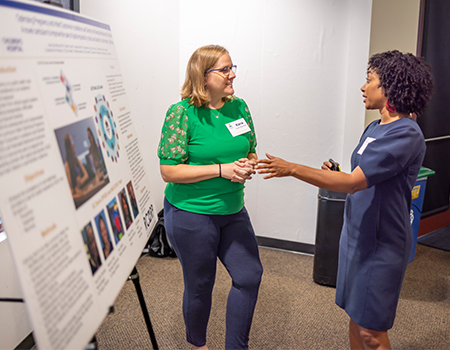 A woman in a green top and a woman in a blue dress converse in front of a research poster in a conference room