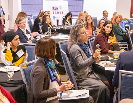 A woman speaks into a microphone surrounded by about 20 people seated around tables in a conference room 