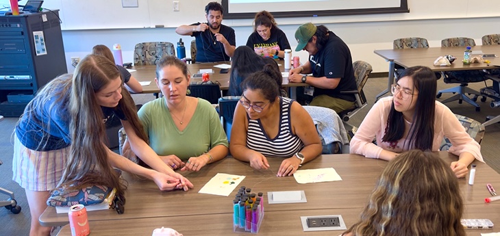 A student leader in blue shirt leans into a table to demonstrate beading techniques to four fellow students 