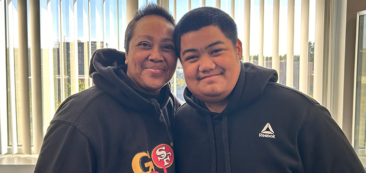 Woman with short hair and black sweatshirt stands next to young man with black sweatshirt.