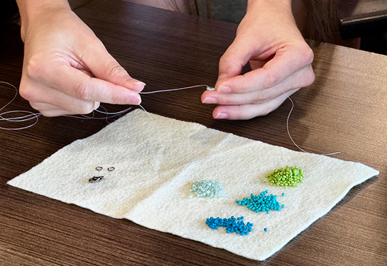 A student’s hands hold thread above four small piles of tiny beads in various colors on a white napkin on a table 