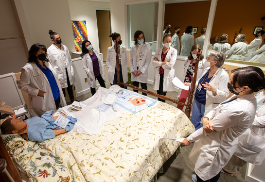 APP fellows wearing white coats and masks surround a bed with manikin laying in it and wound dressings on top of covers