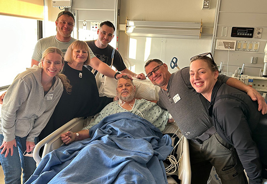 Six smiling family members stand around a hospital bed where an older male patient rests.