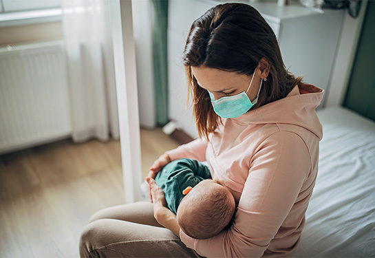 A mother wearing a protective mask breastfeeds her baby son at home.