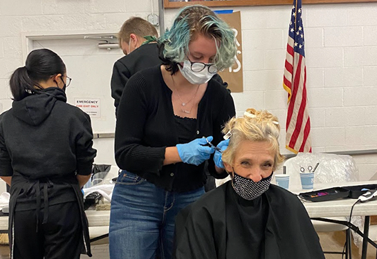 A woman seated with a black beauty salon cape gets her hair cut from a hairdresser standing behind her