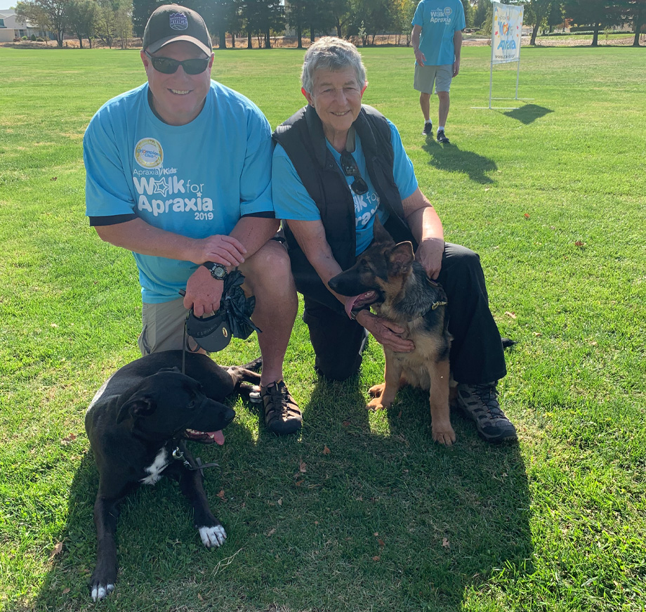 Man and woman kneeling on grass side by side with two dogs laying on ground in front of them