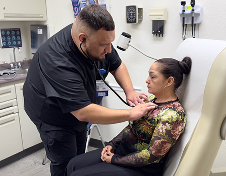 A medical student in black scrubs leans into a medical exam chair to check the heartbeat of a woman who is sitting down