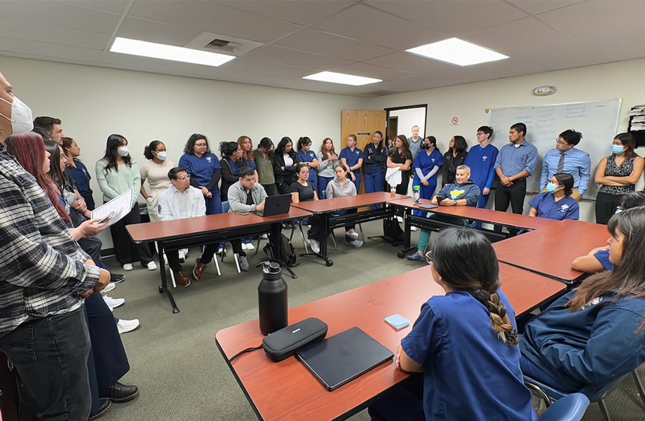 About 40 students wearing blue scrubs and their mentors sit and stand around long tables in a conference room 