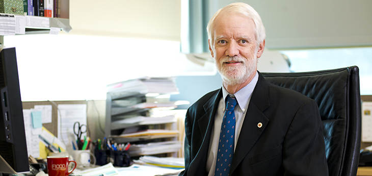 A man with whitish-gray hair sits behind a desk in an office. 