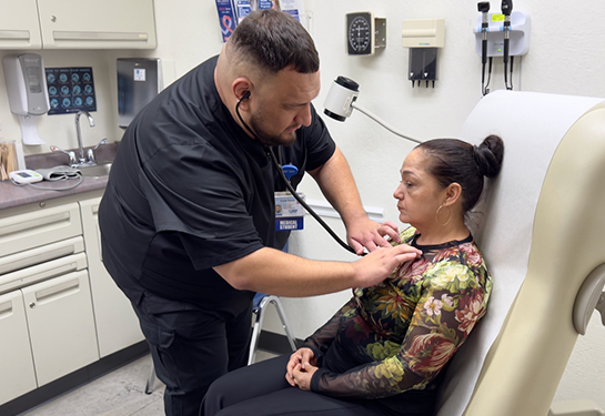 A medical student in black scrubs leans into a medical exam chair to check the heartbeat of a woman who is sitting down