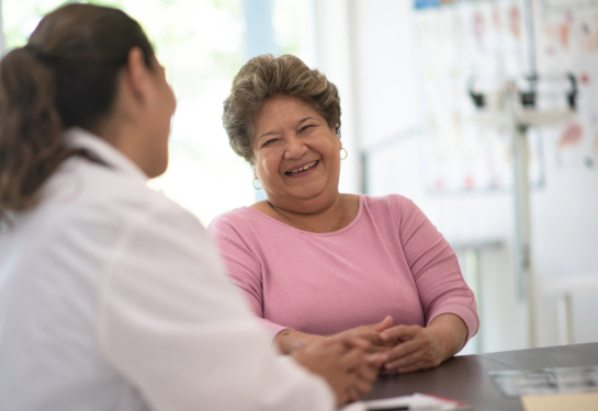 Woman sitting at table facing camera and laughing while speaking to a doctor, whose back of head is shown