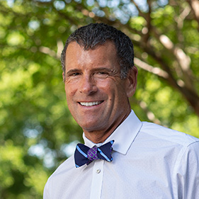 Man in a white shirt and a blue bow tie standing outside with trees in the background.