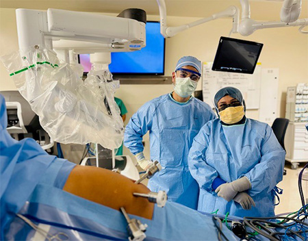 Two people stand side by side wearing blue scrubs, surgery caps, gloves and masks in the operating room.