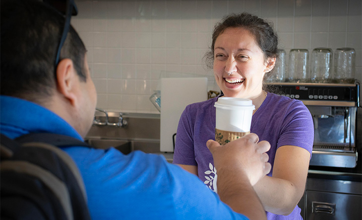 A female café employee in a purple T-shirt hands over a travel coffee cup to a customer while smiling broadly.