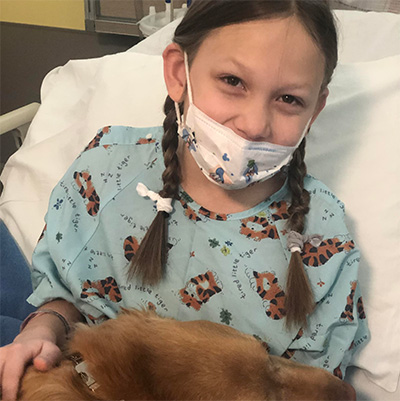 A child in a blue hospital gown with a mask on her face pets a dog.