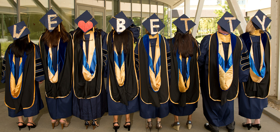 A row of students wearing graduation caps and gowns with backs to camera and mortar boards that read, “I heart Betty”