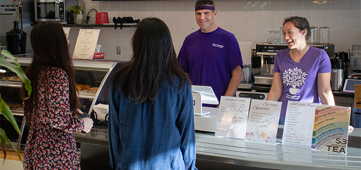 Two customers place their orders at a café. Two employees clad in purple T-shirts chat with them from behind the register. 