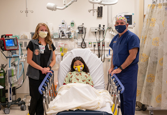 Child laying in hospital bed wearing a face mask and surrounded by a doctor and nurse.